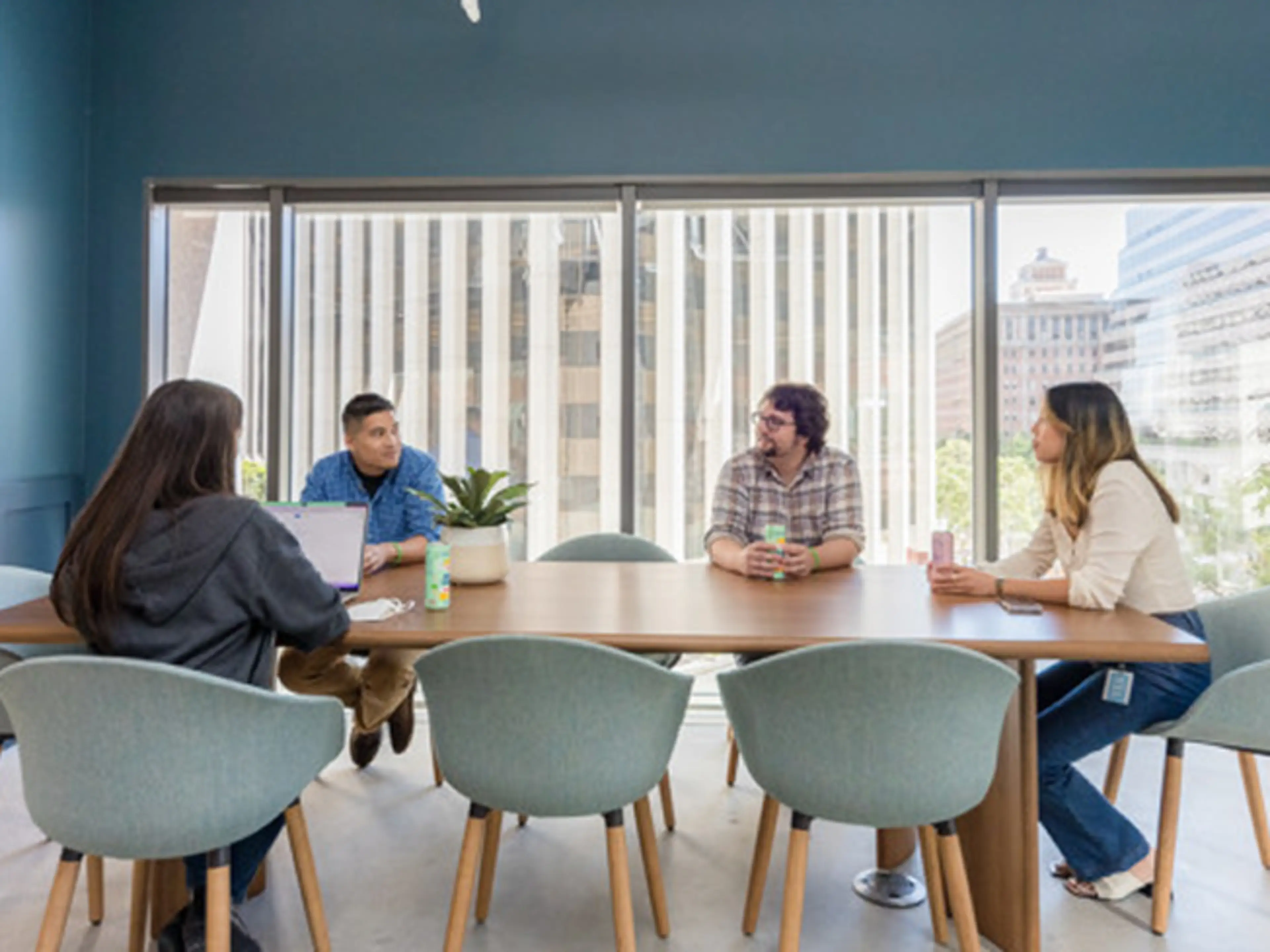 Four people meeting at wooden table in modern blue conference room with large windows overlooking city buildings.